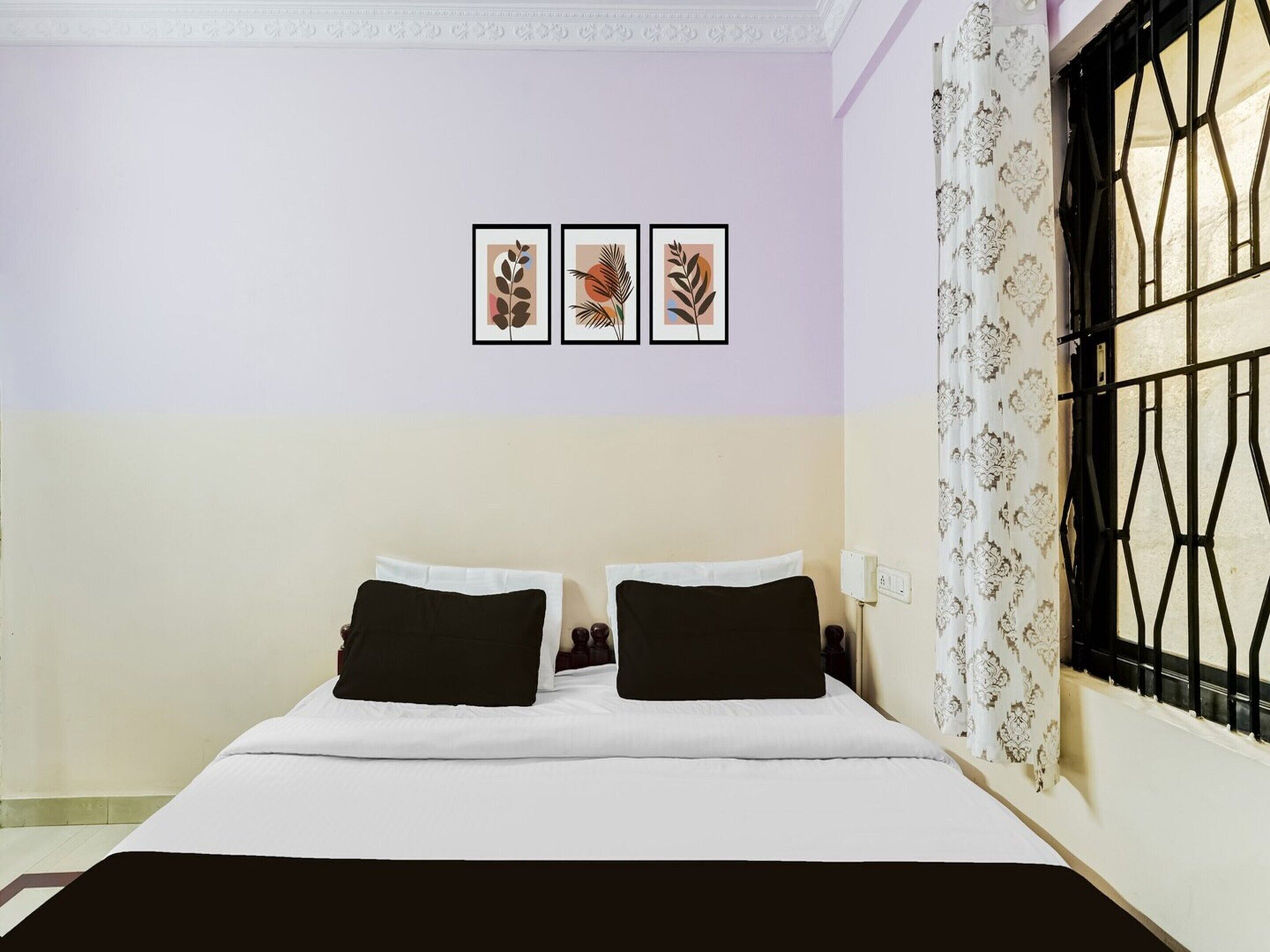 Une chambre à coucher avec un lit, deux oreillers noirs, un couvre-lit blanc et trois motifs de feuilles encadrés accrochés au mur.
