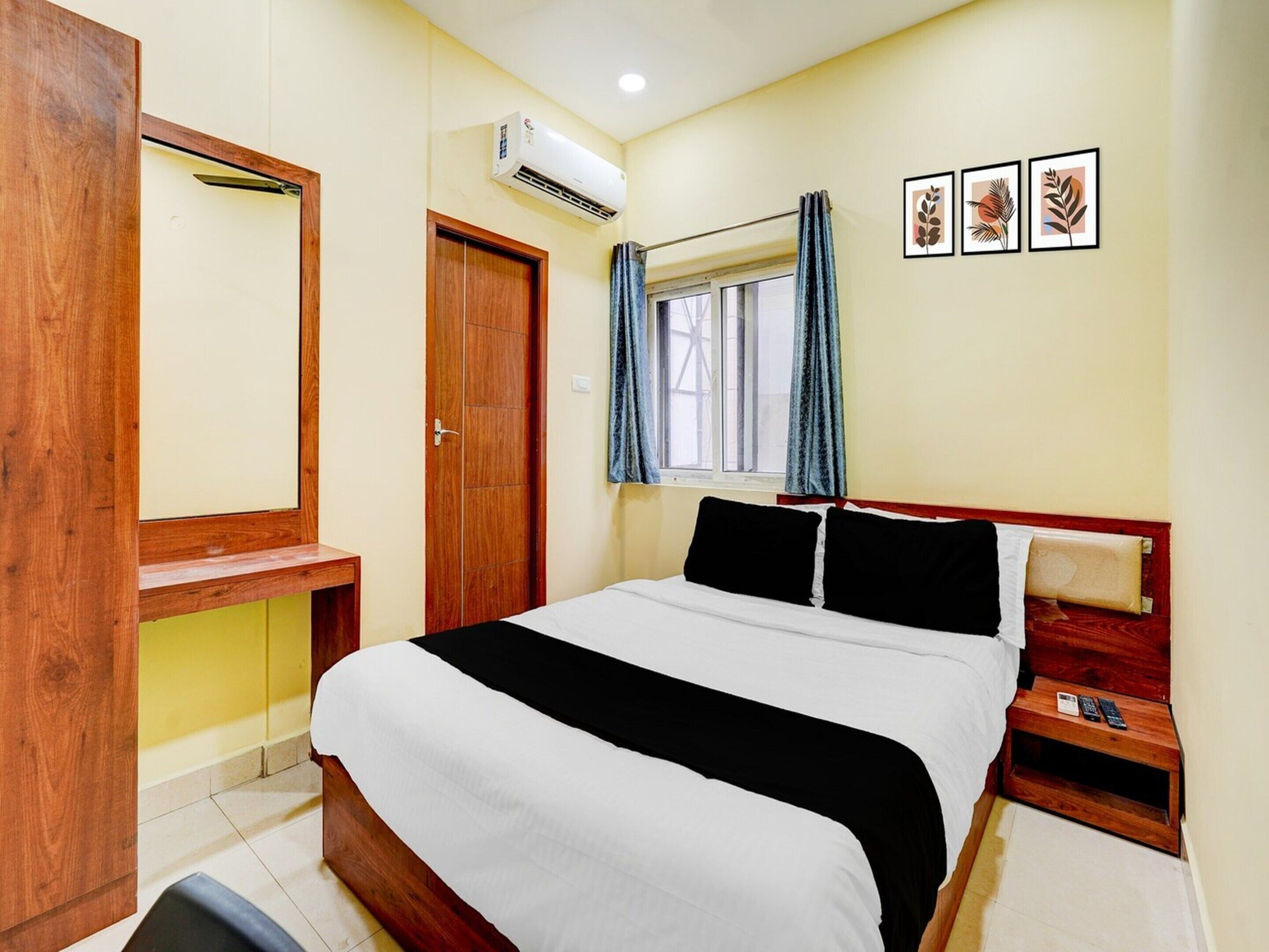 Una habitación de hotel con una cama, un escritorio de madera, aire acondicionado, una ventana con cortinas y tres cuadros en la pared.