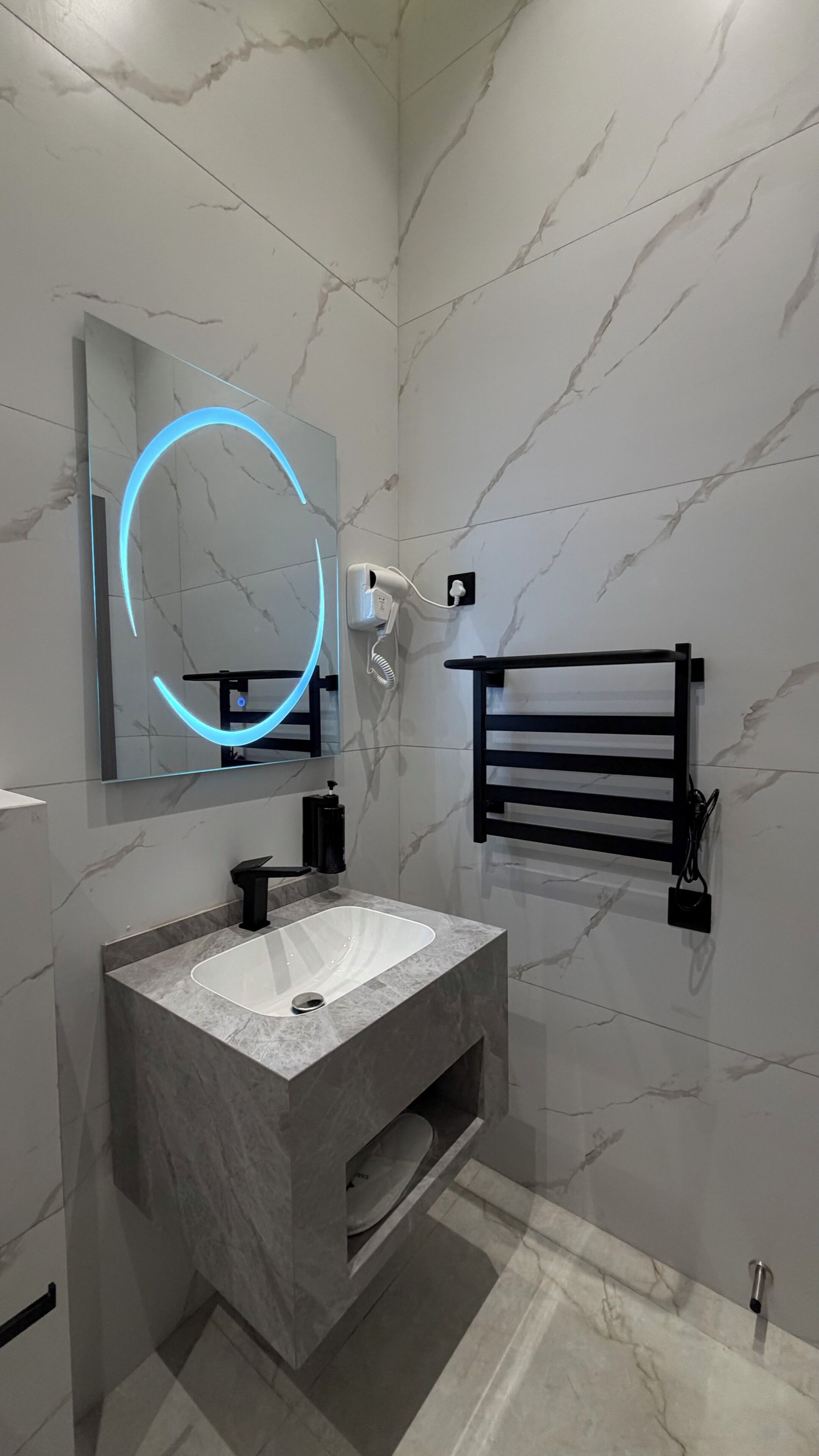 Een moderne badkamer met een marmeren wastafel, een spiegel met blauw licht, een handdoekhouder en een haardroger.