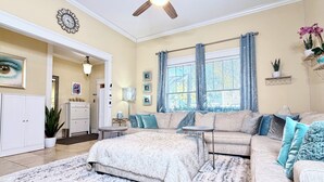Amplia sala de estar con una cama grande, un sofá y un ventilador de techo. Se ve una ventana con cortinas y una planta sobre una estantería.
