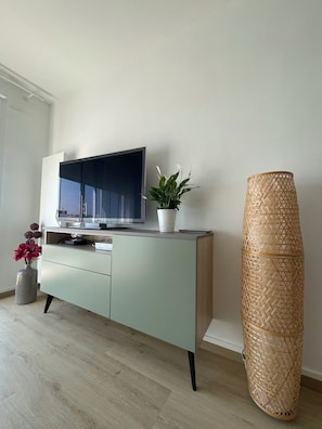 Een moderne woonkamer met een flatscreen televisie op een lichtgekleurd kastje, een potplant en een grote geweven vloerlamp.