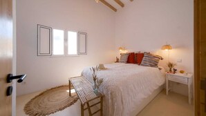 A bedroom with a bed, bedside lamps, a bedside table, a woven bench, and a door. - Duck House (Comporta)