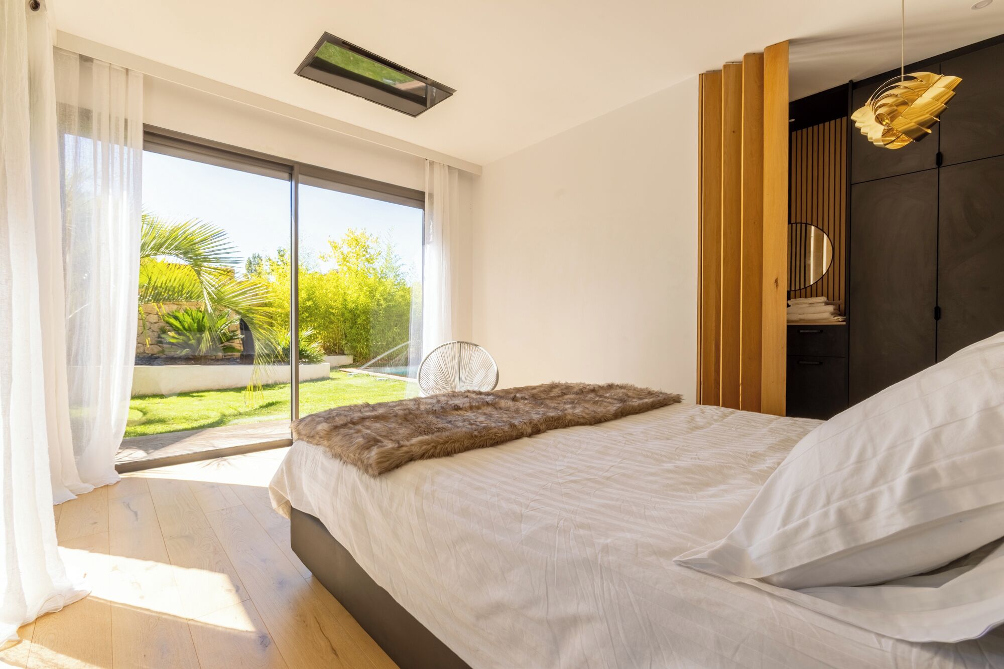 A modern bedroom with a large bed, a view of a garden, and a ceiling fan.