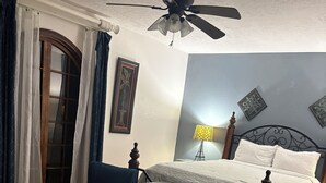 Ein Schlafzimmer mit einem Bett, zwei blauen Stühlen, einem Schreibtisch, einem Deckenventilator, einer Lampe und einem gemusterten Teppich.