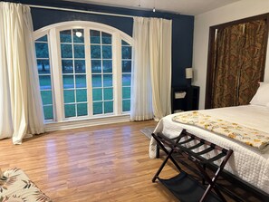 A bedroom with a large window, a bed with a floral bedspread, a wooden floor, and a dark-colored headboard.