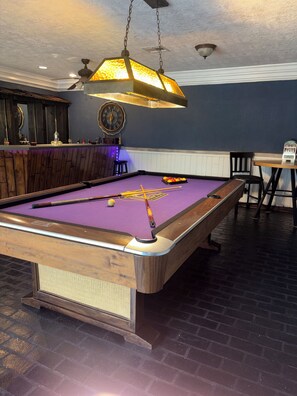 Une table de billard recouverte d’un feutre violet, dotée d’un cadre en bois et surmontée d’un luminaire suspendu.
