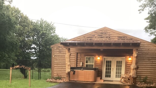 Une cabane rustique en rondins de bois, dotée d’un bain à remous et d’une enseigne en bois sur laquelle on peut lire : « BIENVENUE CHEZ VOUS ».