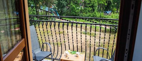 Een balkon met een tafel en stoelen, met uitzicht op een grasveld met bomen en een blauwe constructie in de verte.