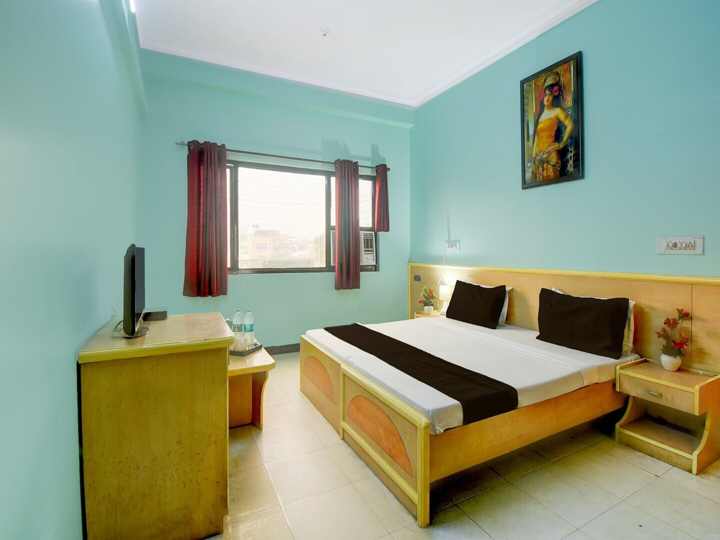 Habitación de hotel con cama, mesita de noche, televisión, ventana con cortinas y un cuadro en la pared.