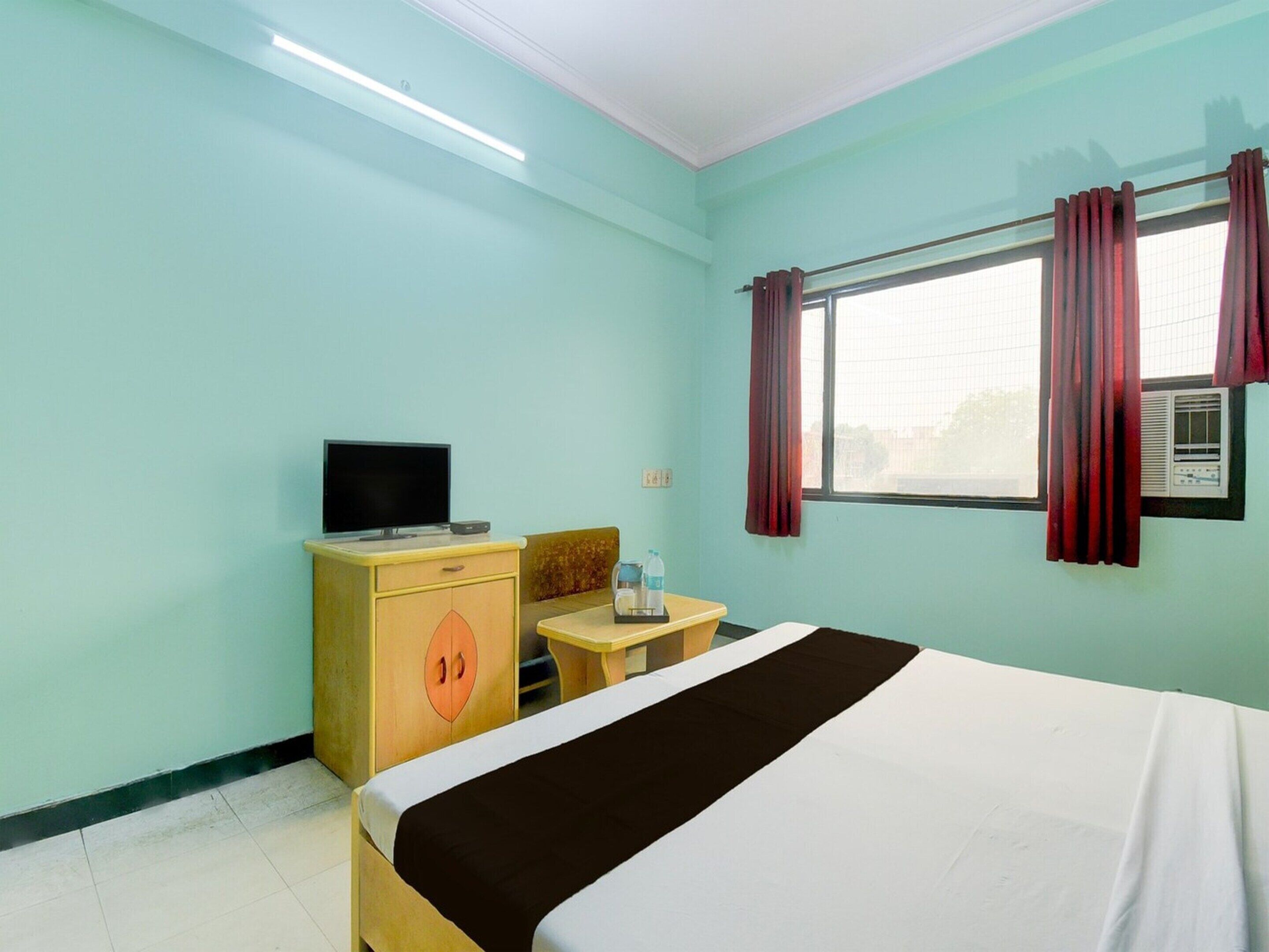 Une chambre d’hôtel avec un lit, un téléviseur posé sur une commode en bois, une petite table avec une bouteille, et une fenêtre avec des rideaux.