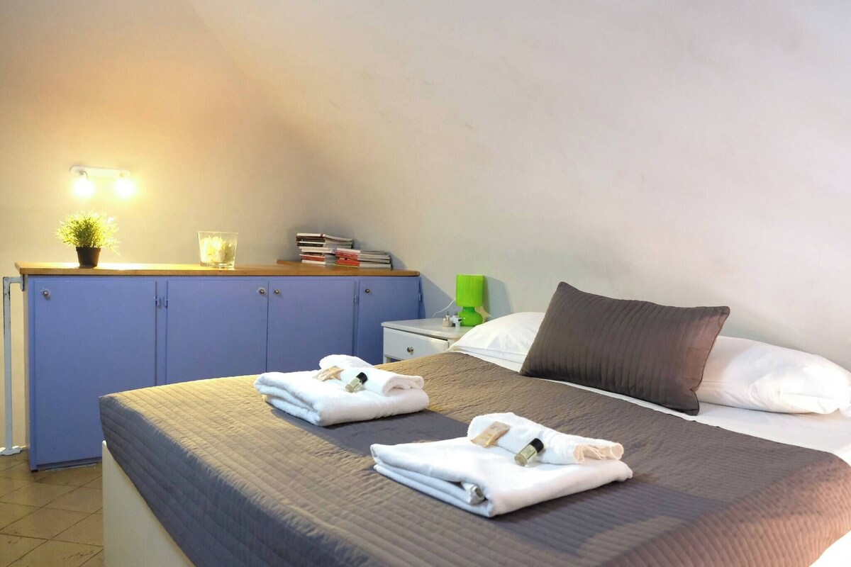 A hotel room with a bed, a nightstand, a lamp, a shelf with books and a plant, and a wall-mounted light.