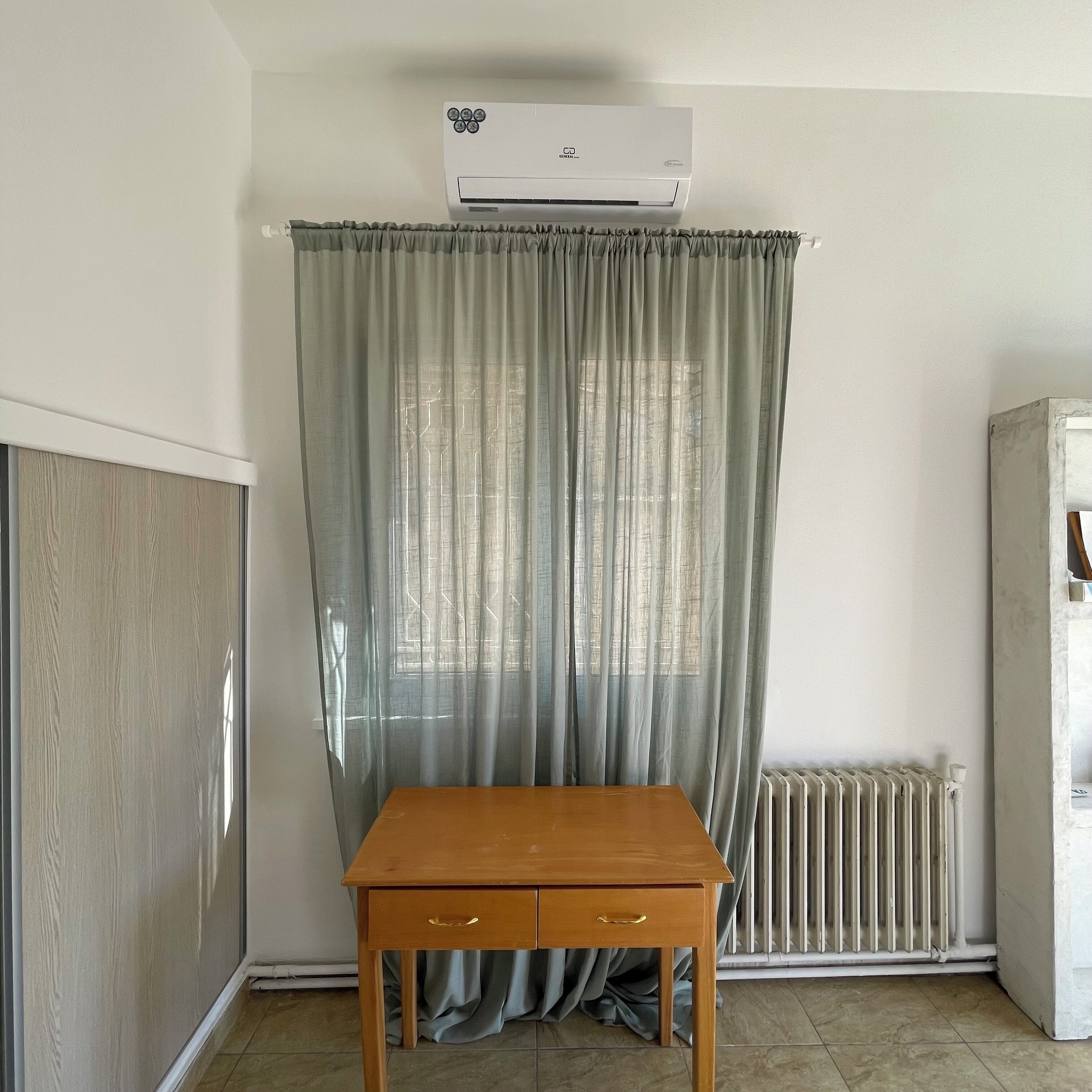 Um quarto com uma mesa de madeira, um radiador e uma janela com cortinas leves.