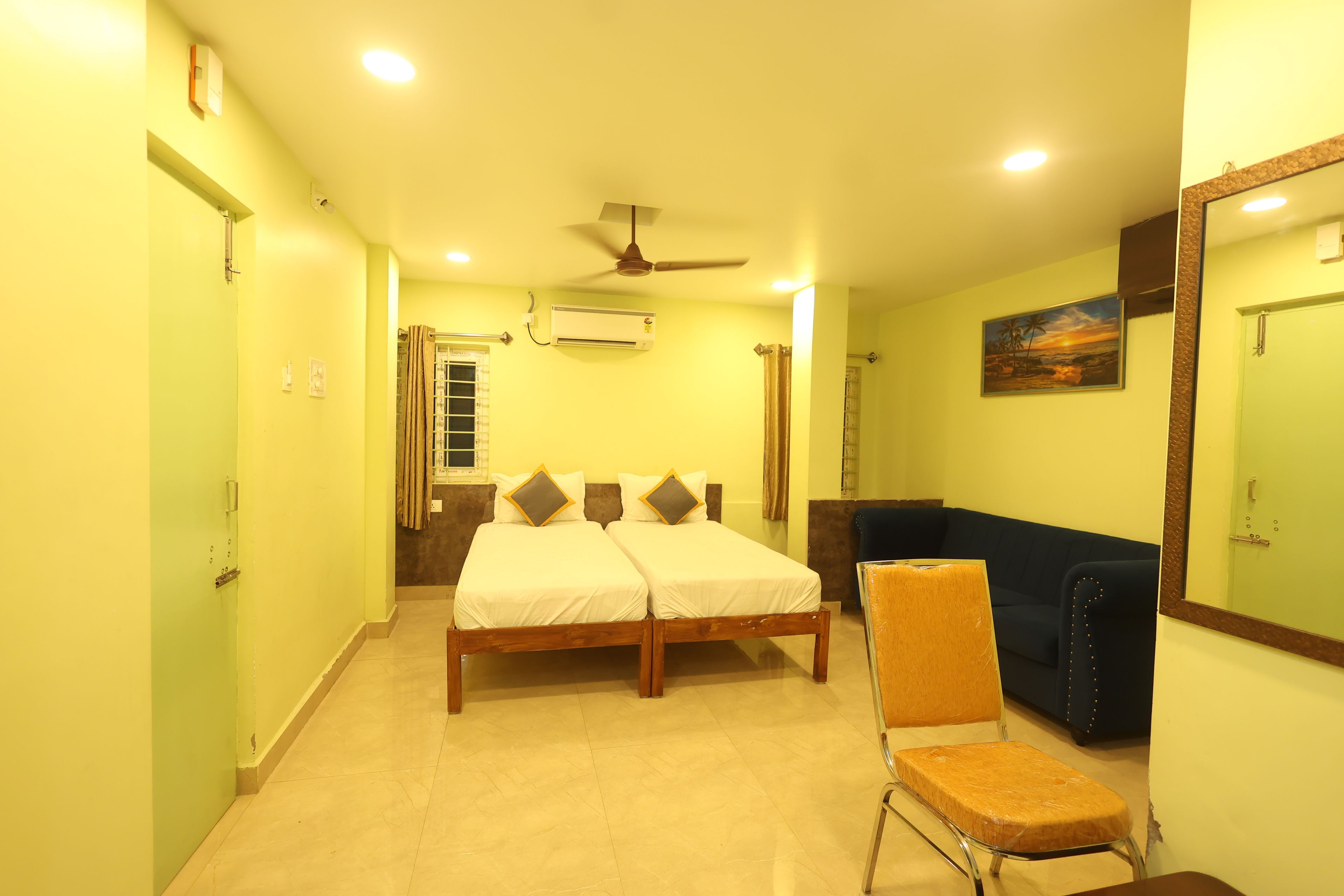 A hotel room with a bed, a seating area, a ceiling fan, an air conditioning unit, and a painting on the wall.