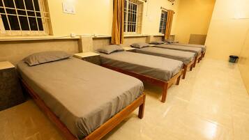 Varias camas de madera con ropa de cama gris en una habitación con paredes amarillas y suelo de baldosas.