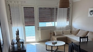 Una sala de estar con sofá, mesa de centro y sillas, ventanas amplias con persianas y una lámpara colgante.