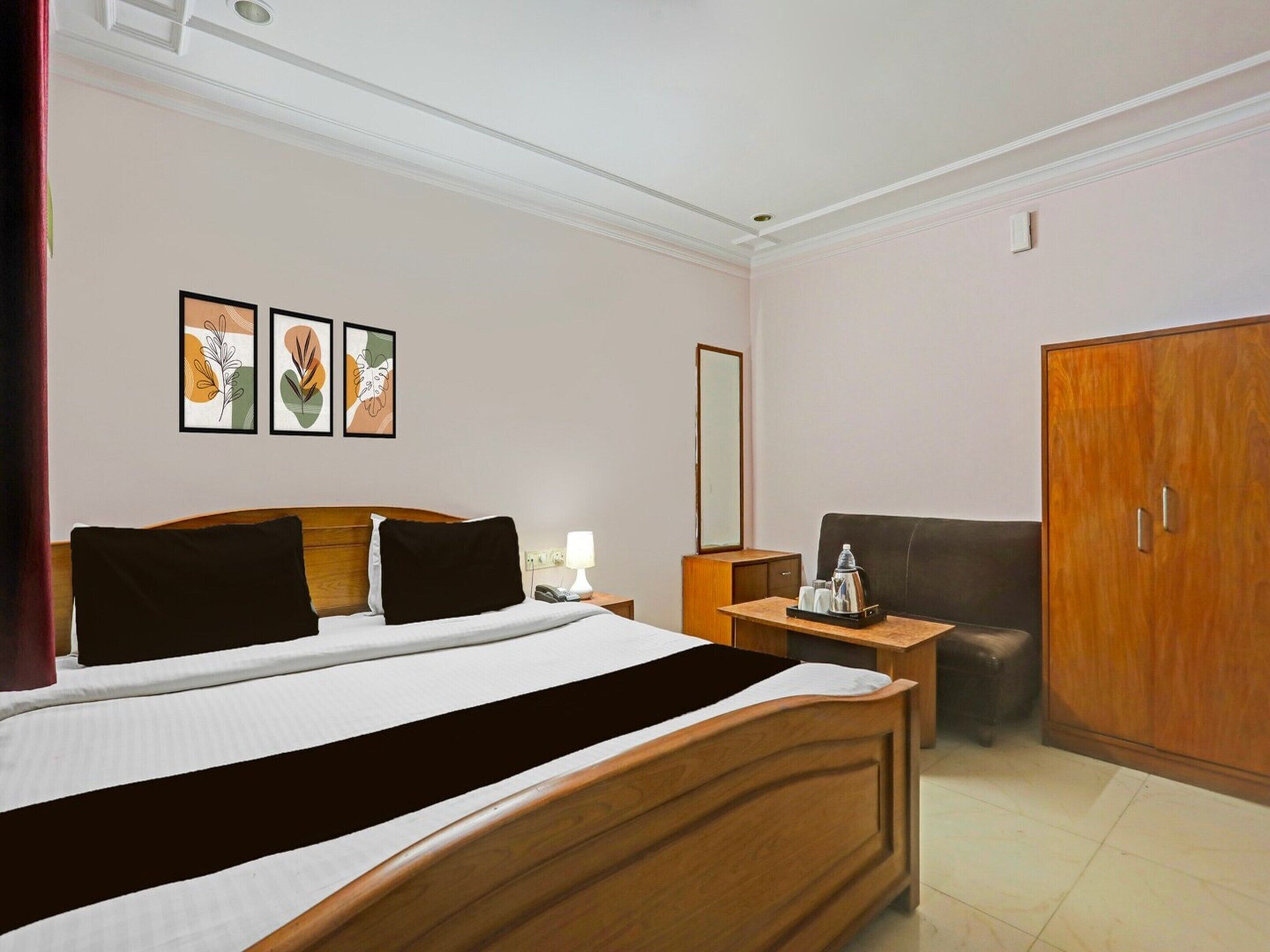A hotel room with a wooden bed, a desk with a lamp, a wardrobe, and three framed artworks on the wall.