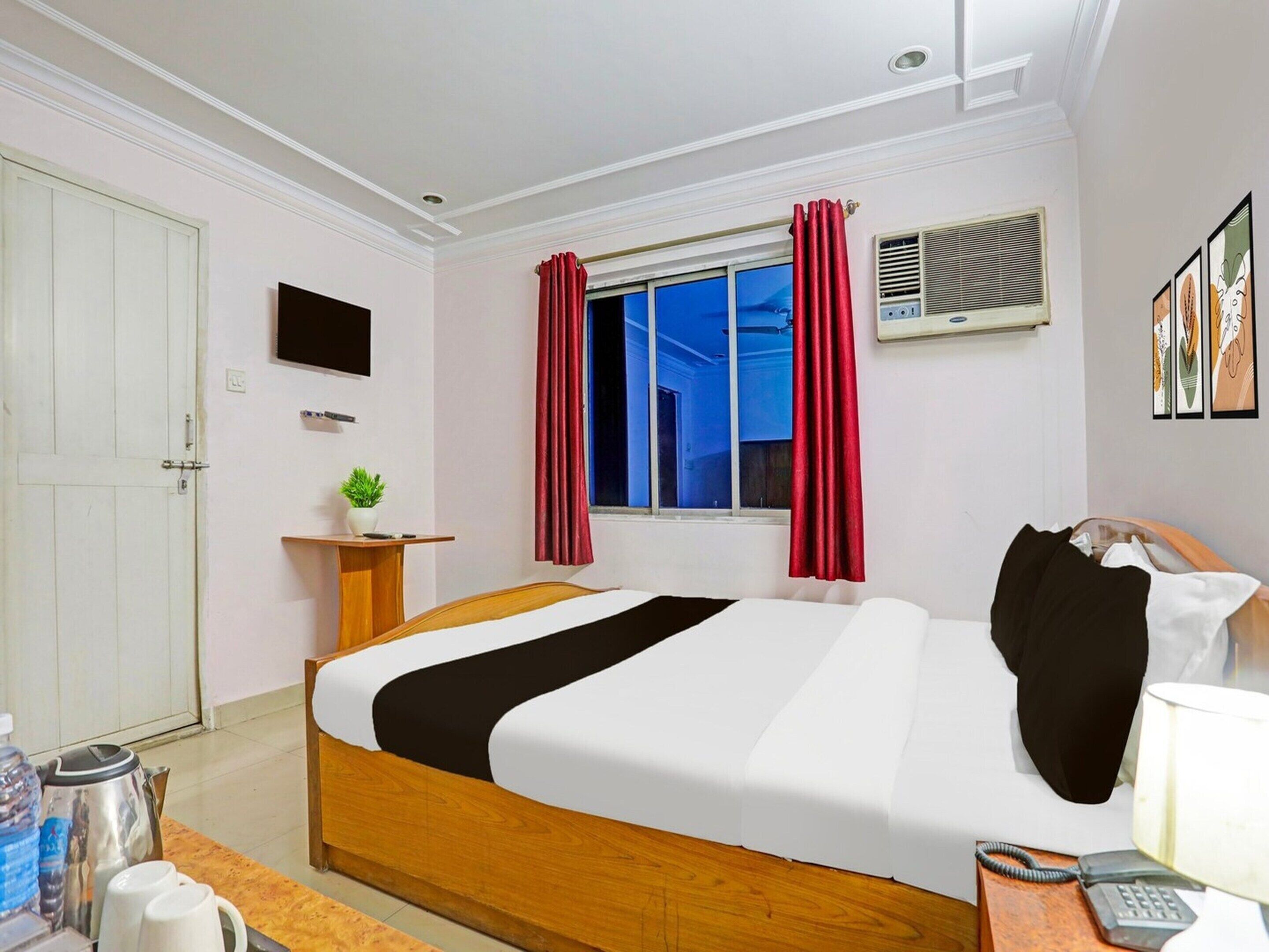 A hotel room with a bed, a desk with a telephone, a window with curtains, a wall-mounted TV, and a wall-mounted air conditioner.