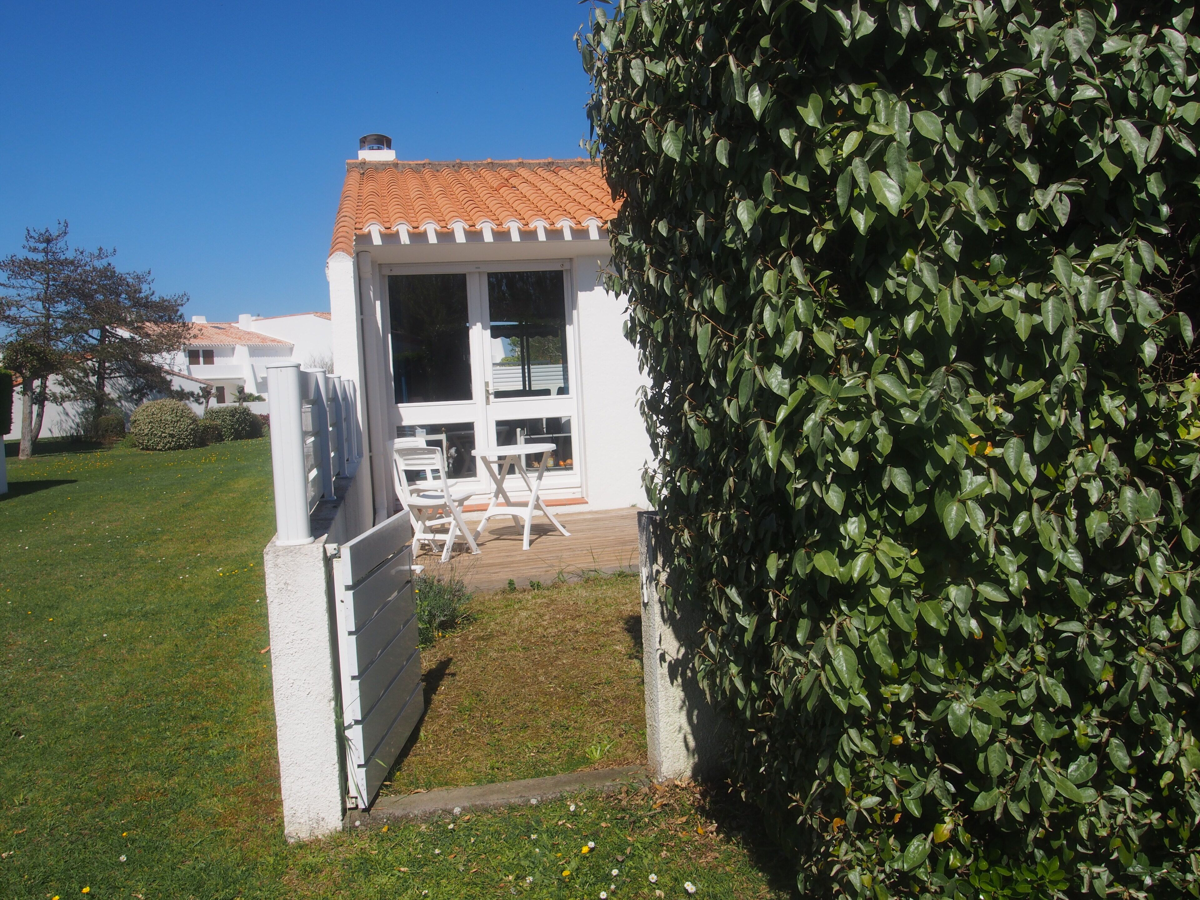 A white building with a tiled roof, a fenced patio area with outdoor furniture, and a well-maintained hedge.