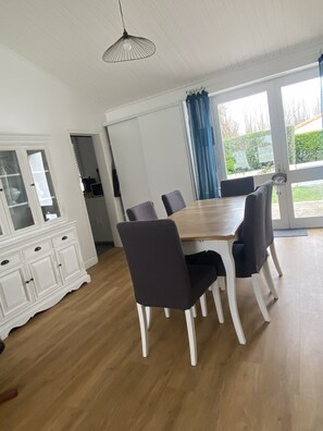 A dining area with a wooden table and chairs, a white cabinet, and a large window with curtains. - Quiet house near the ocean (Saint-Hilaire-de-Riez)