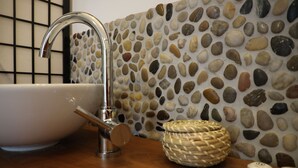 Ett badrum med en vit tvättställ, en modern blandare, en vägg täckt med stenmosaik och en flätad korg på bänkskivan.