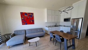 Een moderne leefruimte met een grijze bank, een witte salontafel, een eethoek met stoelen, een keuken met witte kasten, en een groot abstract rood schilderij aan de muur.