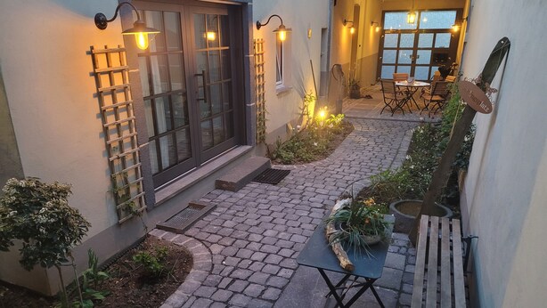 A courtyard with a paved walkway, a wooden bench, potted plants, and a table with chairs.