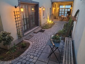 Une cour avec un chemin pavé, un banc en bois, des plantes en pot et une table avec des chaises.