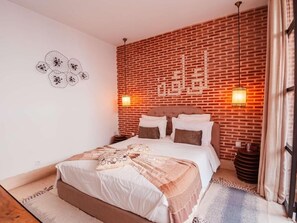 Un dormitorio con pared de ladrillo, una cama con ropa de cama blanca, dos luces fijadas a la pared y un adorno decorativo en la pared.
