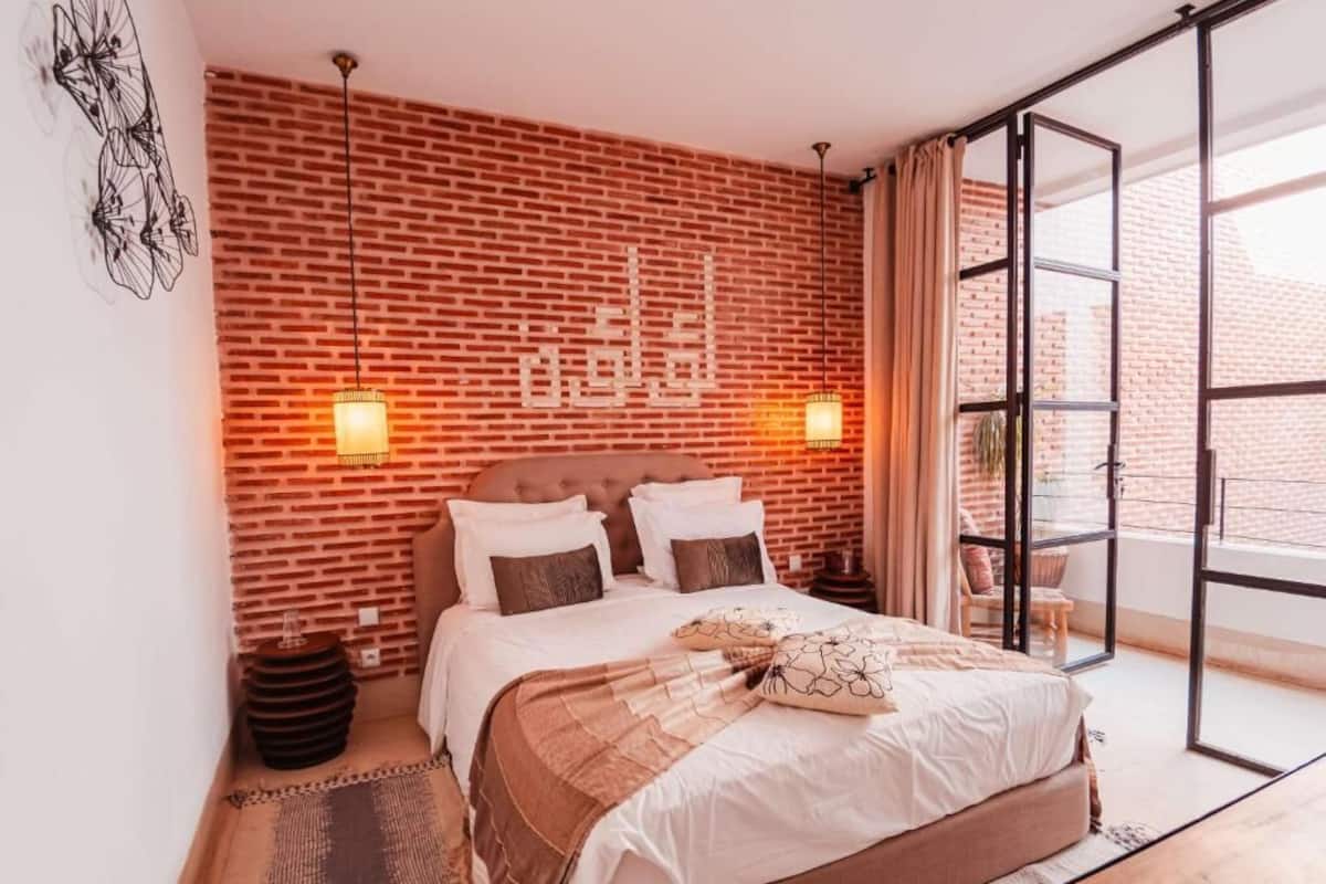 Een slaapkamer met een bakstenen muur, een bed met meerdere kussens, twee wandlampen, een klein rond tafeltje en een groot raam met gordijnen.