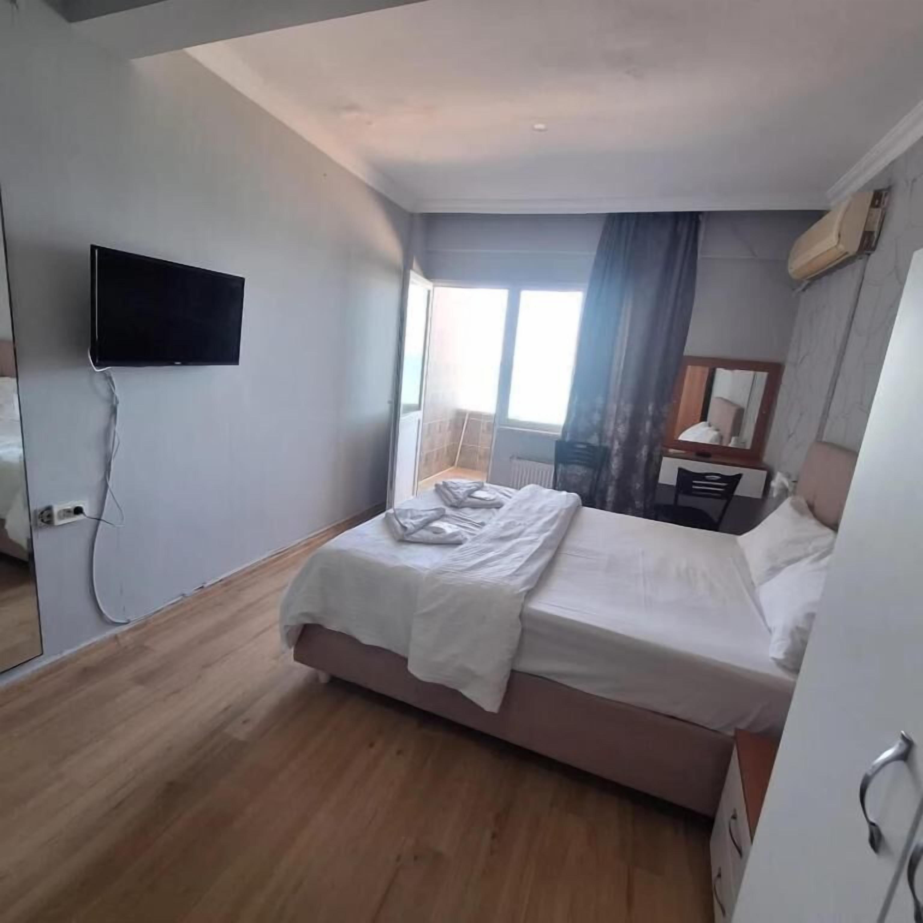 Habitación de hotel con cama, televisor de pantalla plana montado en la pared, ventana con cortinas y mesita de noche con lámpara.