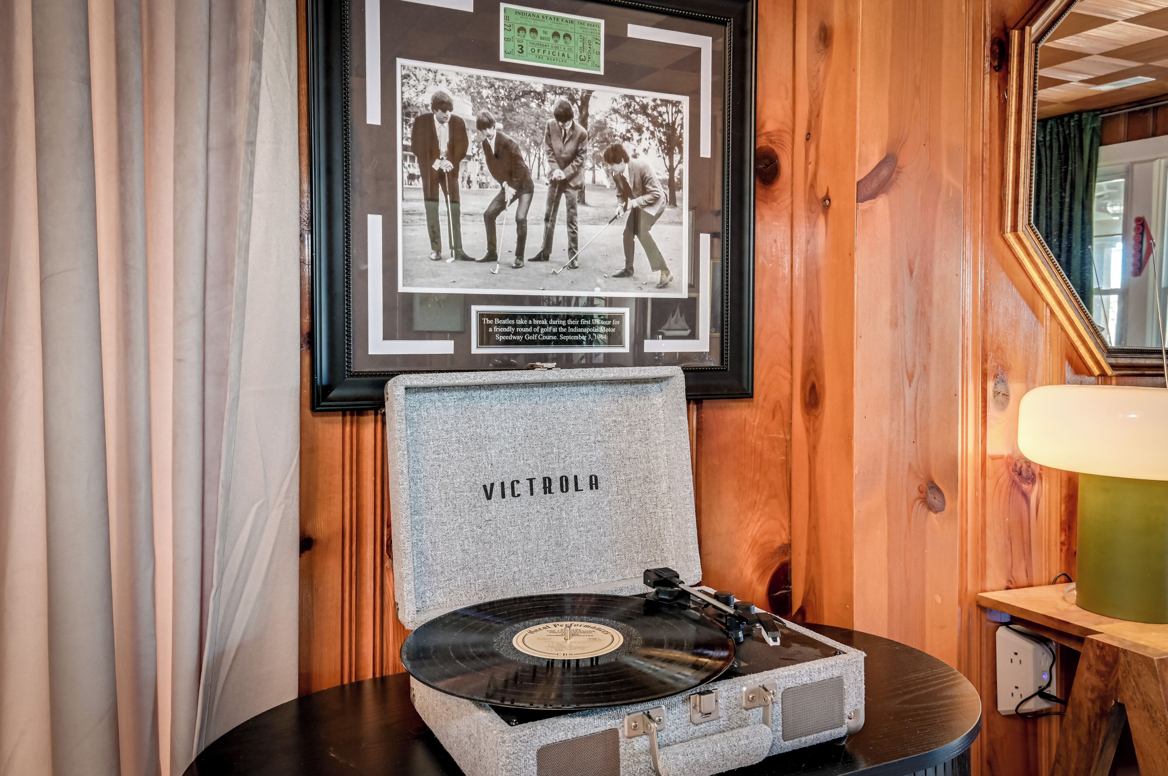Une photographie encadrée des Beatles sur un terrain de golf, une platine vinyle avec un disque, et un miroir reflétant une pièce aux murs en bois.