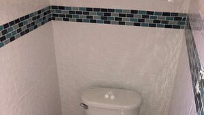 A toilet with a white seat and lid, a blue sticker on the seat, and a corner with white and teal mosaic tiles. - Cozy 1-bedroom bungalow in charming Pleasant Harbour, Morgan’s Bluff with AC (Morgan’s Bluff)
