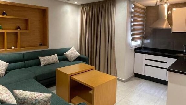 Ein modernes Wohnzimmer mit einer Eckcouch, einem Holztisch und einer Küchenzeile mit weißen Schränken und einer schwarzen Arbeitsplatte.