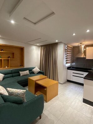 A modern living room with a sectional sofa, a wooden coffee table, and a kitchen area with white cabinets and a black countertop. - Hôtel Résidence Ramsis (Nador)