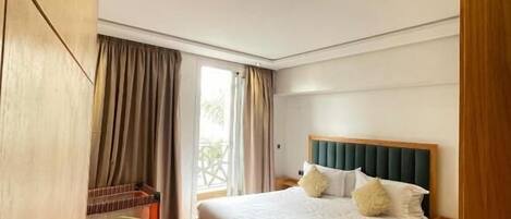 Ein modernes Schlafzimmer mit einem großen Bett, Holzboden, einem grünen Sessel und Blick ins Freie durch das Fenster.