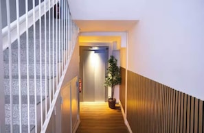 Een moderne hotelgang met een houten vloer, een witte leuning en een plant in pot.