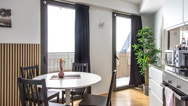Une cuisine moderne équipée d’une cuisinière, d’un four à micro-ondes et de placards, un coin repas avec une table ronde et des chaises, et une grande fenêtre avec des rideaux.