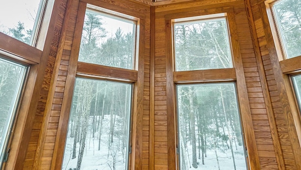 En stuga med träpanel på insidan, ett sluttande tak, stora fönster och utsikt över en snötäckt skog.