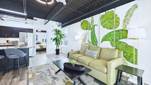 A modern living space with a kitchen, dining area, and a large wall mural of tropical leaves. - Stay Park Cruise Modern Loft by Port -unit 200 (Cape Canaveral)
