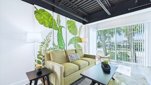 A modern living room with a sofa, a coffee table, and a side table. There is a large window with a view of palm trees and buildings. - Stay Park Cruise Modern Loft by Port -unit 200 (Cape Canaveral)