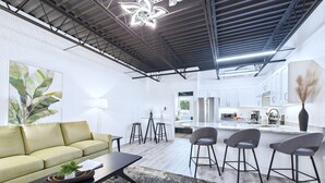 Un salon moderne avec un canapé, une table basse et des tabourets de bar. On y trouve également un coin cuisine avec des meubles blancs et une table à manger.