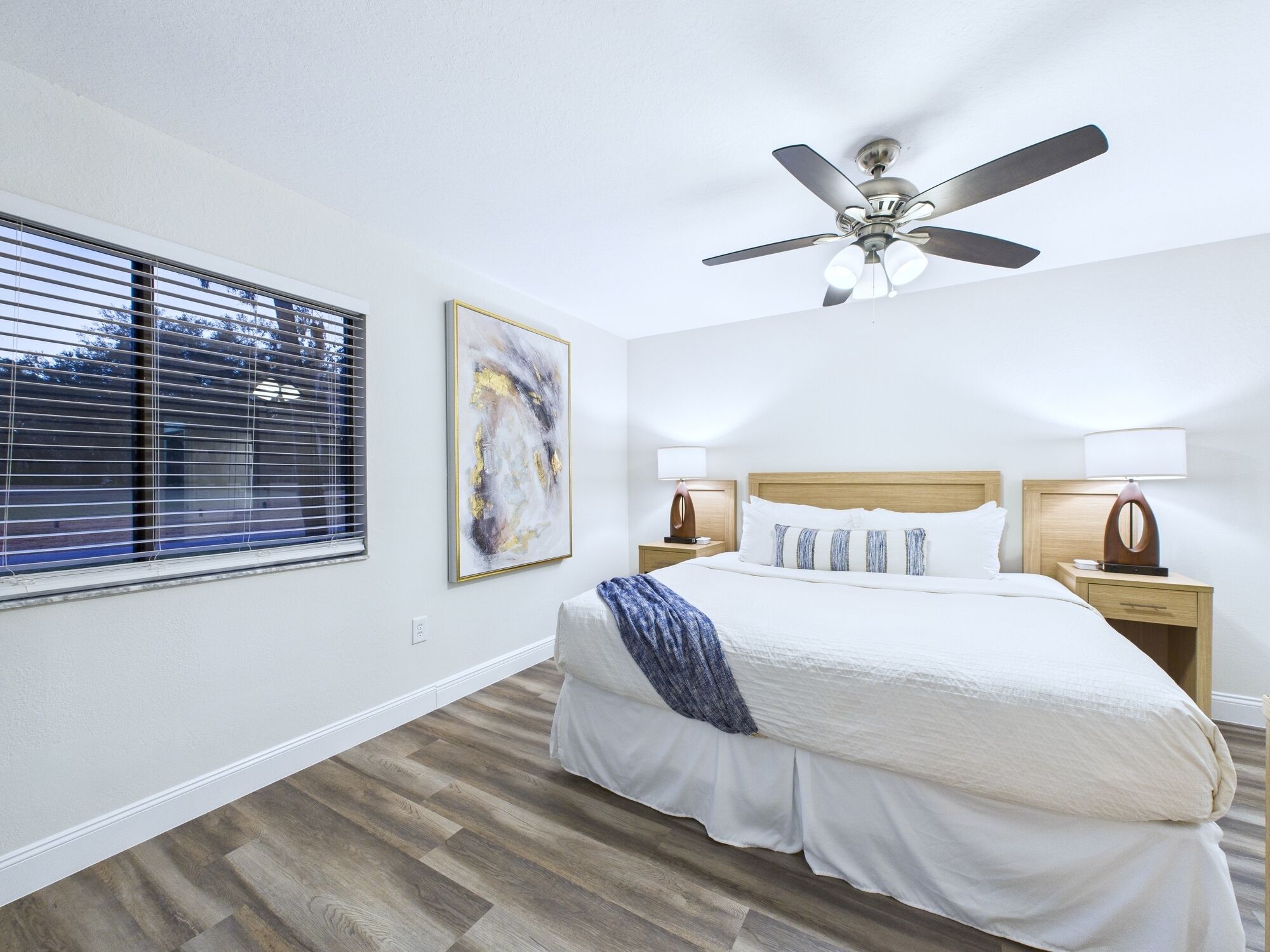 A bedroom with a bed, a ceiling fan, two bedside tables with lamps, a window with blinds, and a painting on the wall.