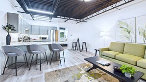 A modern kitchen with grey cabinets, a central island with bar stools, and a dining area with a green sofa and a coffee table. - Stylish Loft Minutes From Beach Port - Unit 204 (Cape Canaveral)