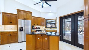 A modern kitchen with wooden cabinets, a stainless steel refrigerator, a black countertop, and a ceiling fan. - Indian Harbor Beach Pool Retreat (Indian Harbour Beach)
