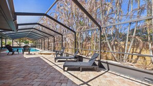 A covered outdoor area with a pool, lounge chairs, and a view of palm trees. - Indian Harbor Beach Pool Retreat (Indian Harbour Beach)