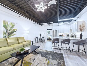 Una sala moderna con sofá, una mesita de centro y sillas, una cocina con armarios blancos, y un techo con vigas a la vista y luminarias.