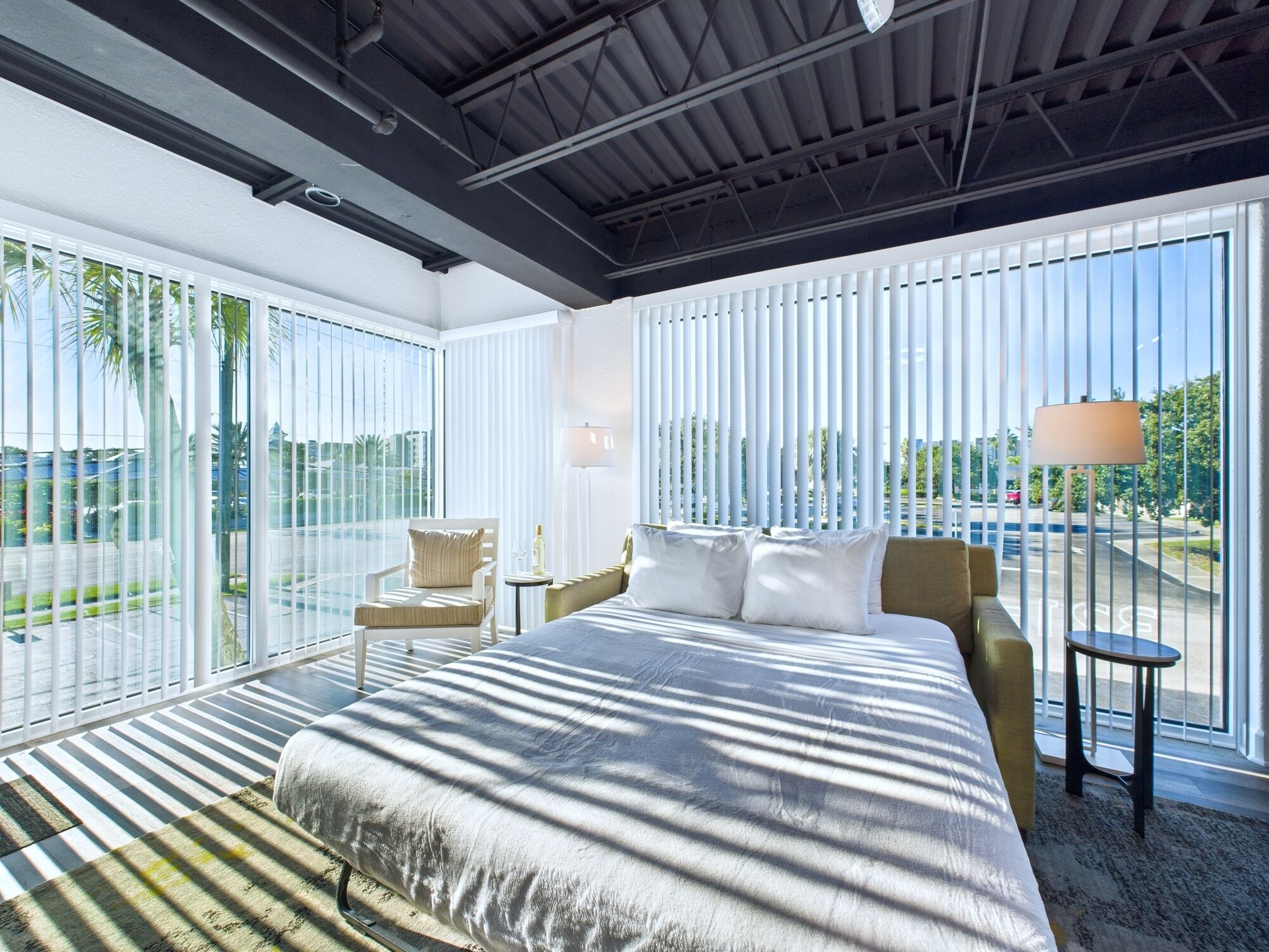 A modern hotel room with a large bed, a chair, a nightstand, and a lamp. The room has large windows with vertical blinds and a view of the outside.