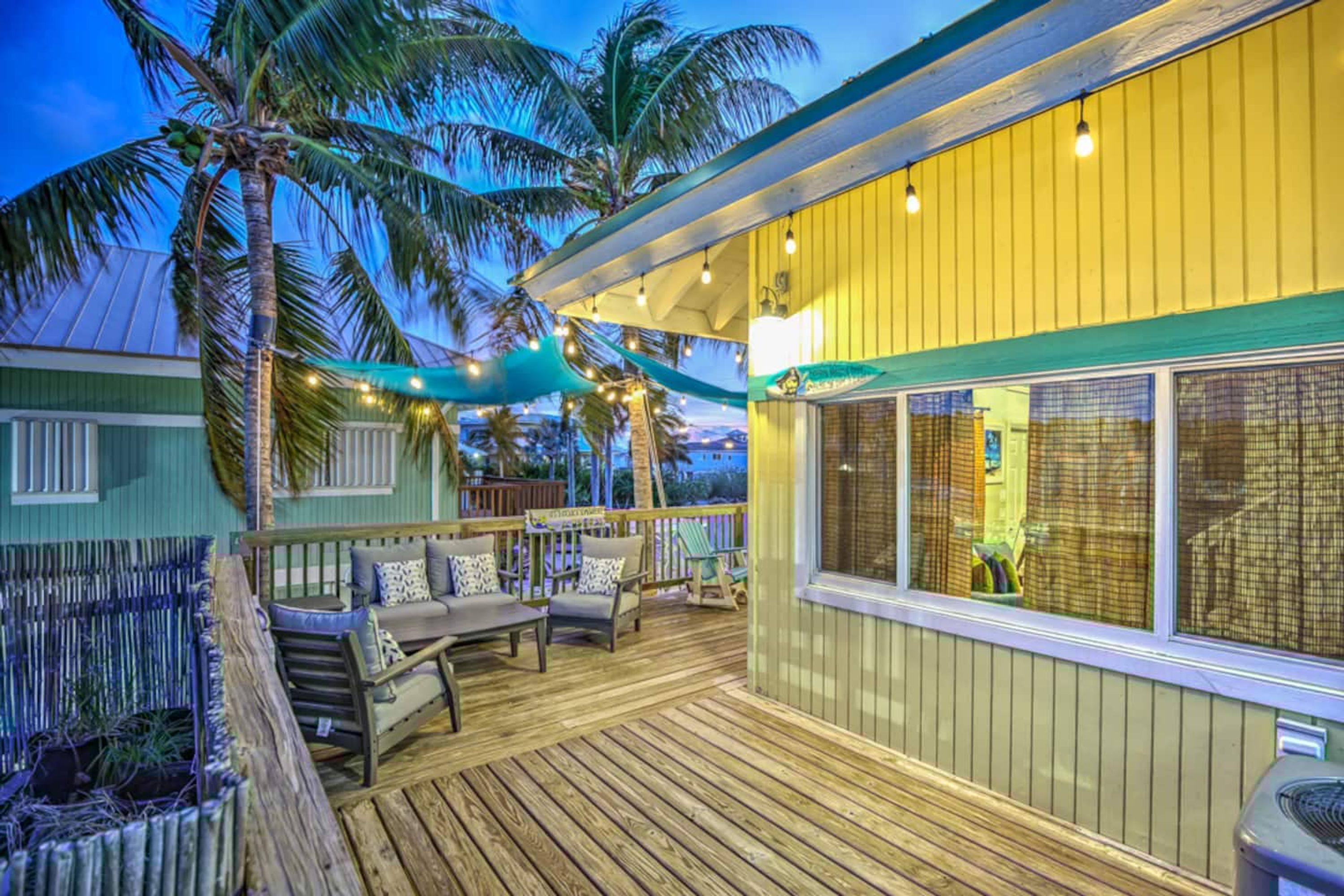 Une terrasse en bois avec des sièges d’extérieur, un bâtiment jaune doté de grandes fenêtres et des palmiers.