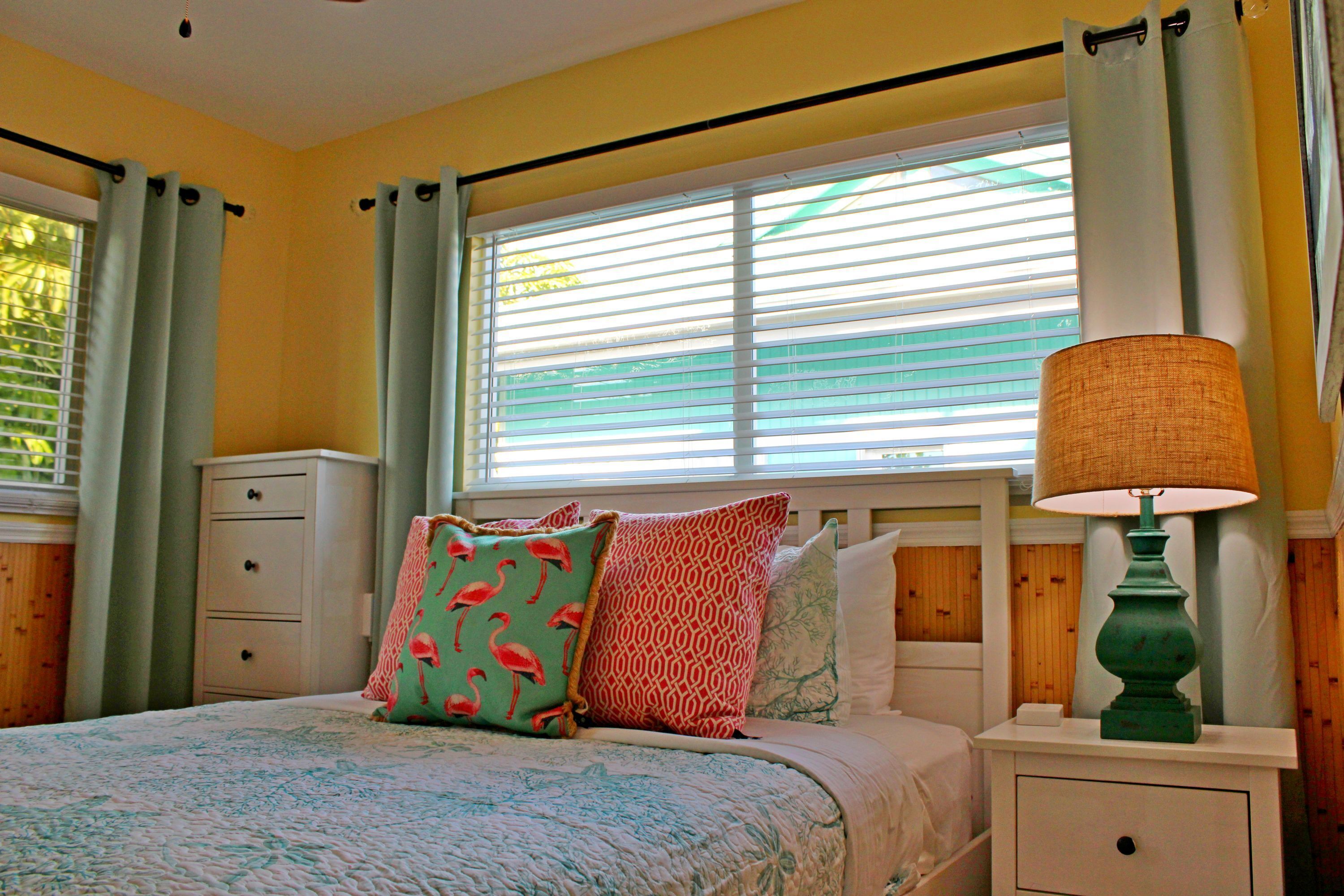 A bedroom with a bed, a nightstand, a lamp, a window with blinds, and a dresser.