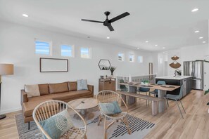A modern living room with a sofa, two wicker chairs, a coffee table, a dining area with a bench, a ceiling fan, and a kitchen with a refrigerator. - Walk to Beach & Trails | Modern, Cozy Retreat (Carolina Beach)